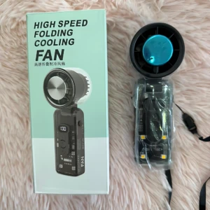 Ventilador portátil plegable negro con pantalla digital, caja de cartón verde claro y accesorios en plástico transparente sobre superficie de peluche rosa.