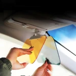 Visera de coche con lentes intercambiables: una en tono ámbar y otra en gris claro, siendo ajustadas por manos frente al parabrisas para bloquear el sol.