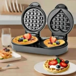 Waflera Sokany 2 en 1 con placas intercambiables, forma cuadrada y corazón, waffles dorados con frutas frescas sobre mesa de madera.