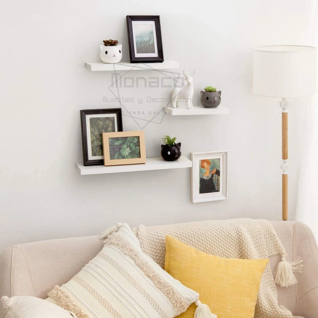 Tres repisas flotantes blancas en pared, exhibiendo marcos de fotos, macetas pequeñas y figuras decorativas, sobre sofá beige con cojines.