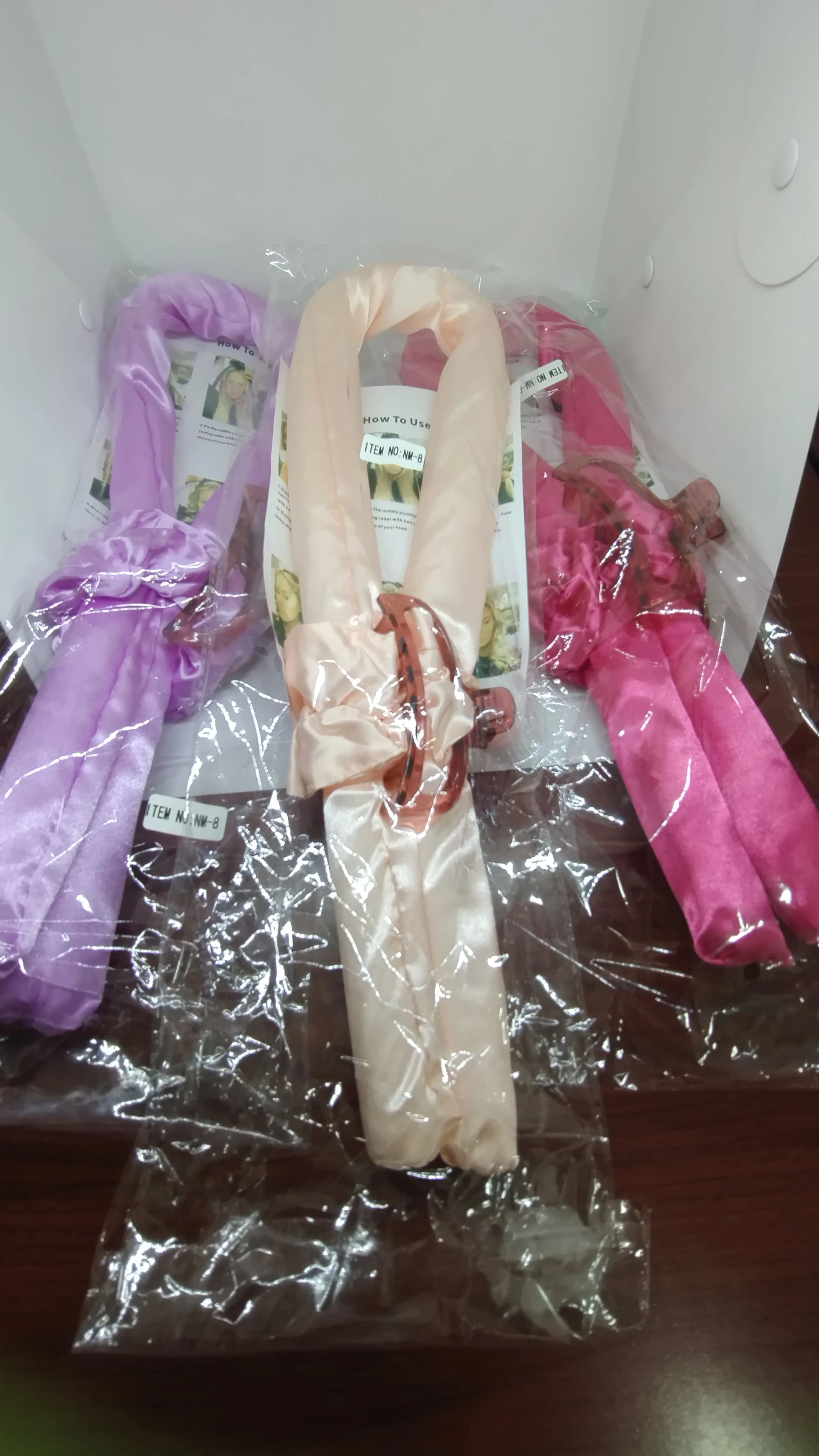 Tres onduladores de pelo enrollados en plástico transparente, en tonos lavanda, crema y fucsia, con diadema incluida.
