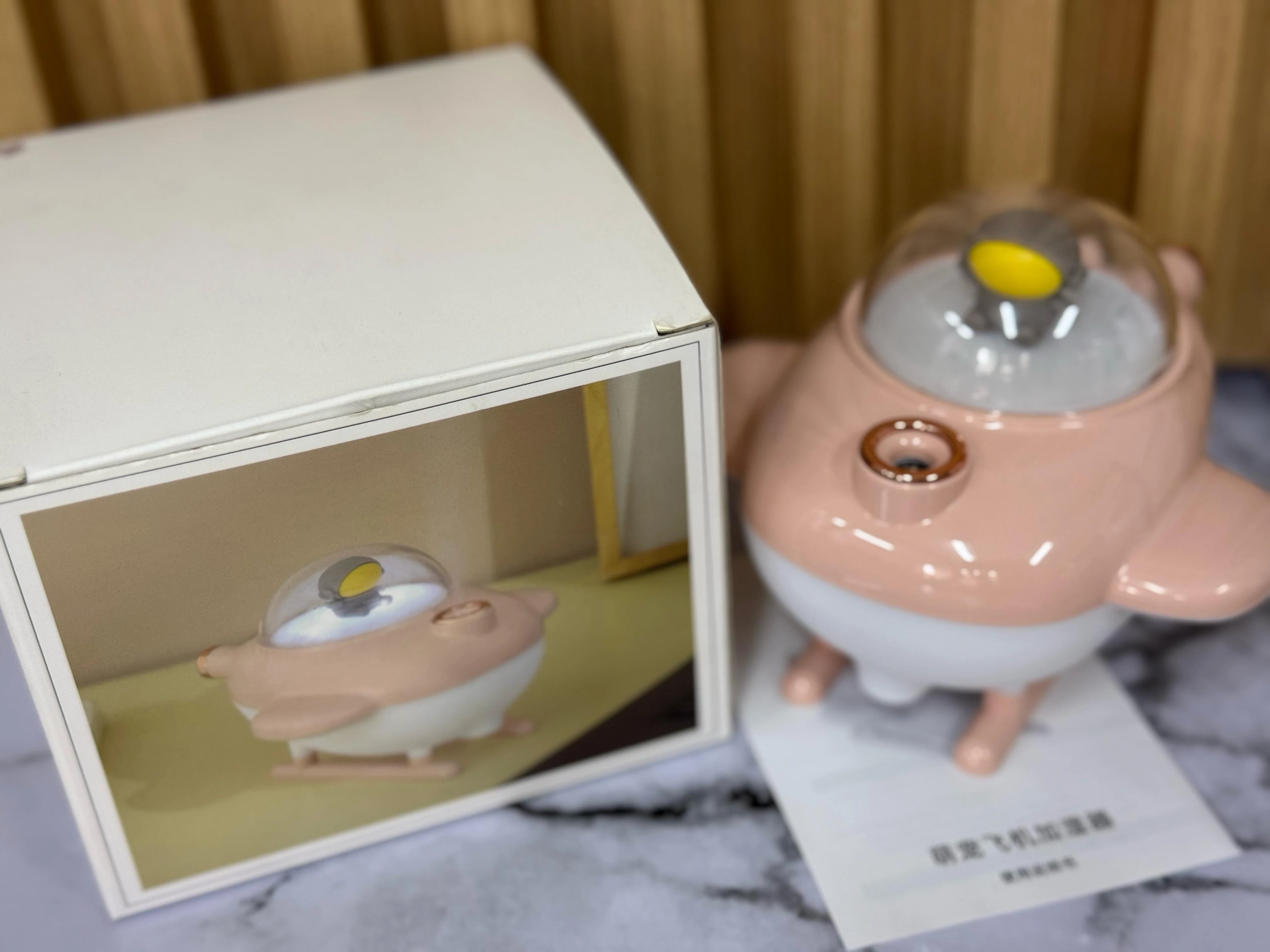 Humificador de aire en forma de nave espacial rosa pastel con cúpula transparente y base blanca, junto a su caja de presentación.
