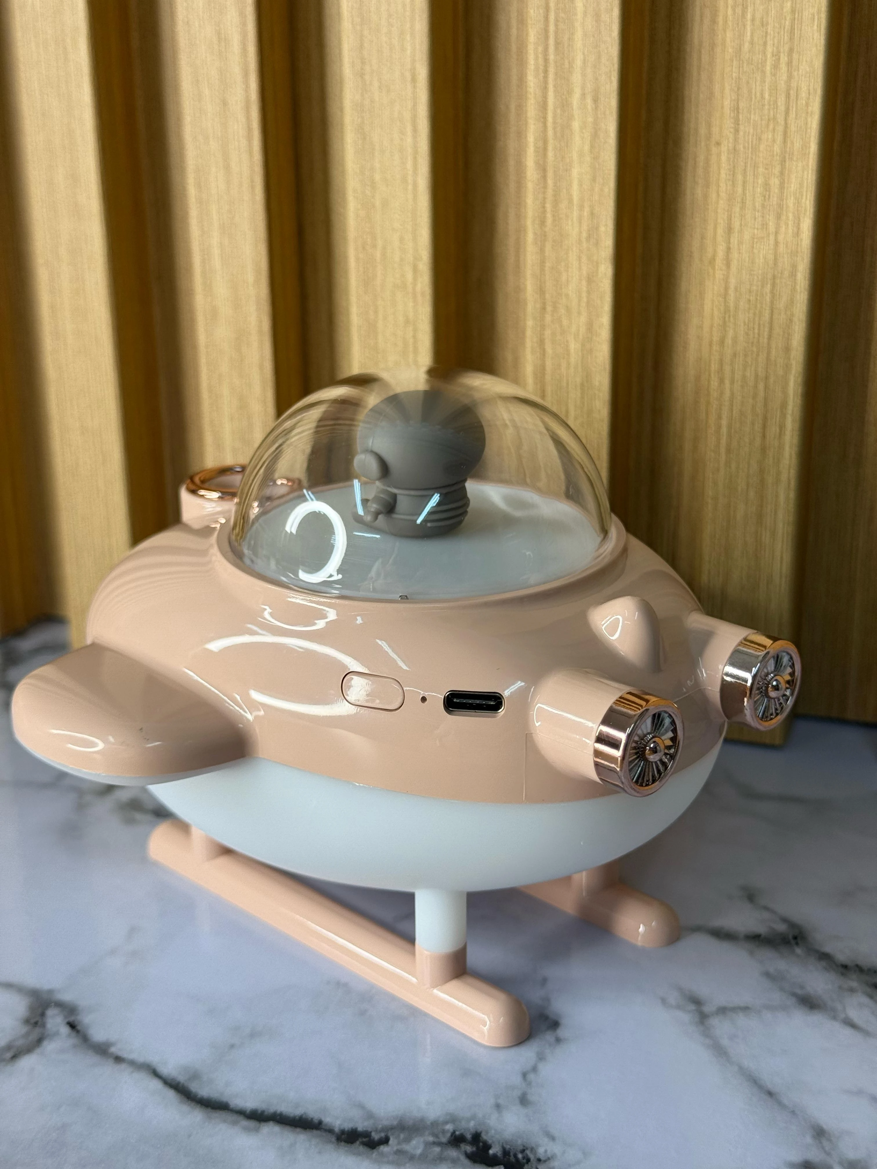 Humificador de aire en forma de nave espacial, color beige y blanco, con cúpula transparente y detalles metálicos, sobre superficie de mármol.