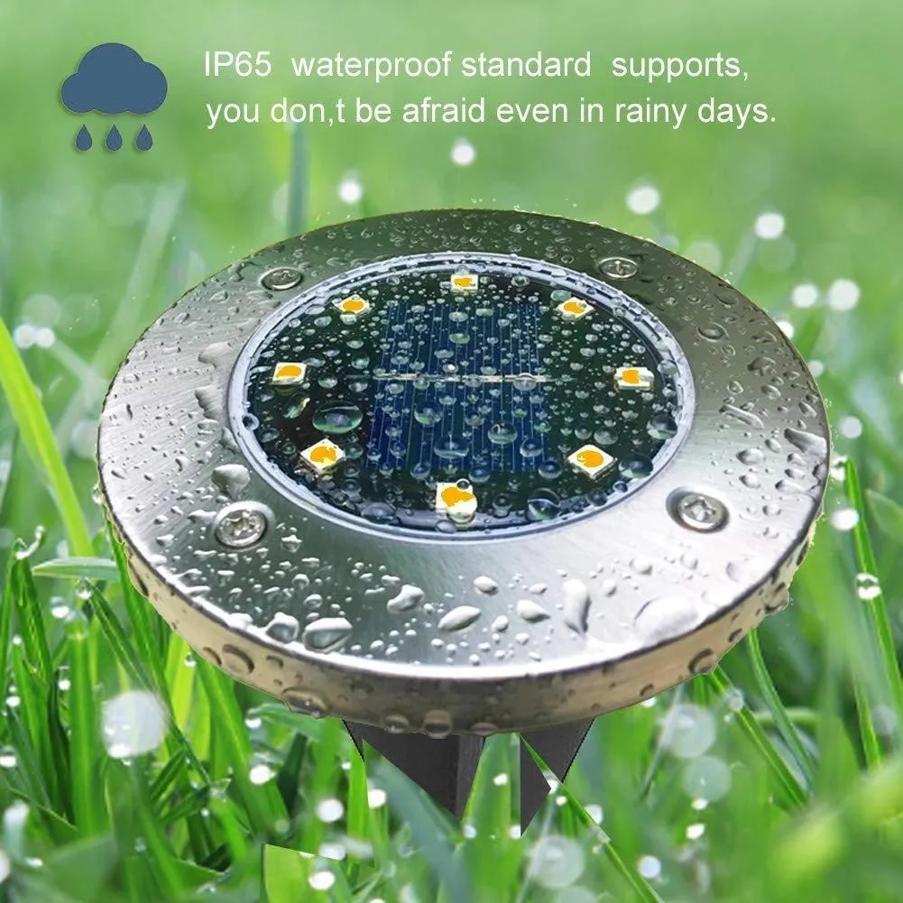 Luz solar de piso IP65 con sensor automático, vista superior con gotas de agua sobre su superficie metálica y LEDs amarillos, instalada en césped verde.