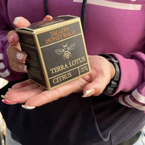 Bálsamo Terra Lotus en caja negra con detalles dorados, mostrando etiqueta frontal con de abeja y texto Tallow Honey Balm. Presentación compacta, manos sosteniendo producto.