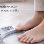 Báscula de baño electrónica cuadrada con pantalla digital mostrando 62.4 kg, pies descalzos sobre superficie de cristal templado, diseño minimalista en tonos plateados y grises.