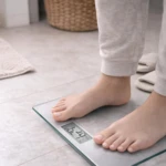 Báscula de baño cuadrada de cristal templado, con pantalla digital mostrando 57.0 kg, usada por persona en pijama sobre piso de cerámica.