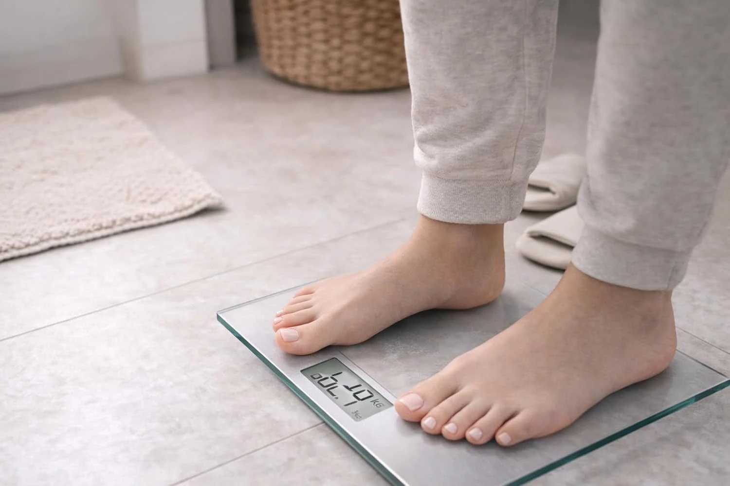 Báscula de baño cuadrada de cristal templado, con pantalla digital mostrando 57.0 kg, usada por persona en pijama sobre piso de cerámica.