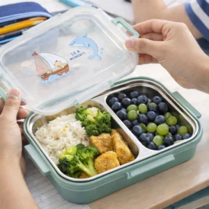 Lonchera escolar de acero inoxidable en color menta, con tapa transparente decorada con dibujos marinos. Contiene arroz, brócoli, nuggets de pollo, uvas verdes y moras, dispuestas en compartimentos separados.