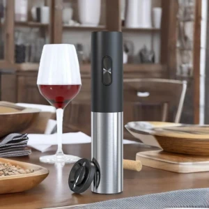 Abrebotellas eléctrico inalámbrico con cuerpo metálico y tapa negra, junto a copa de vino y accesorios sobre mesa de madera.