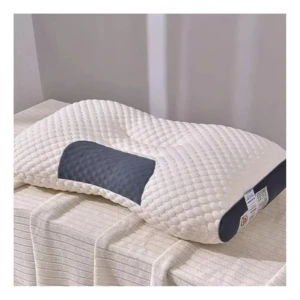 Almohada ortopédica blanca con textura de bolitas y zona cervical acolchada en gris oscuro, sobre cama con sábanas blancas.