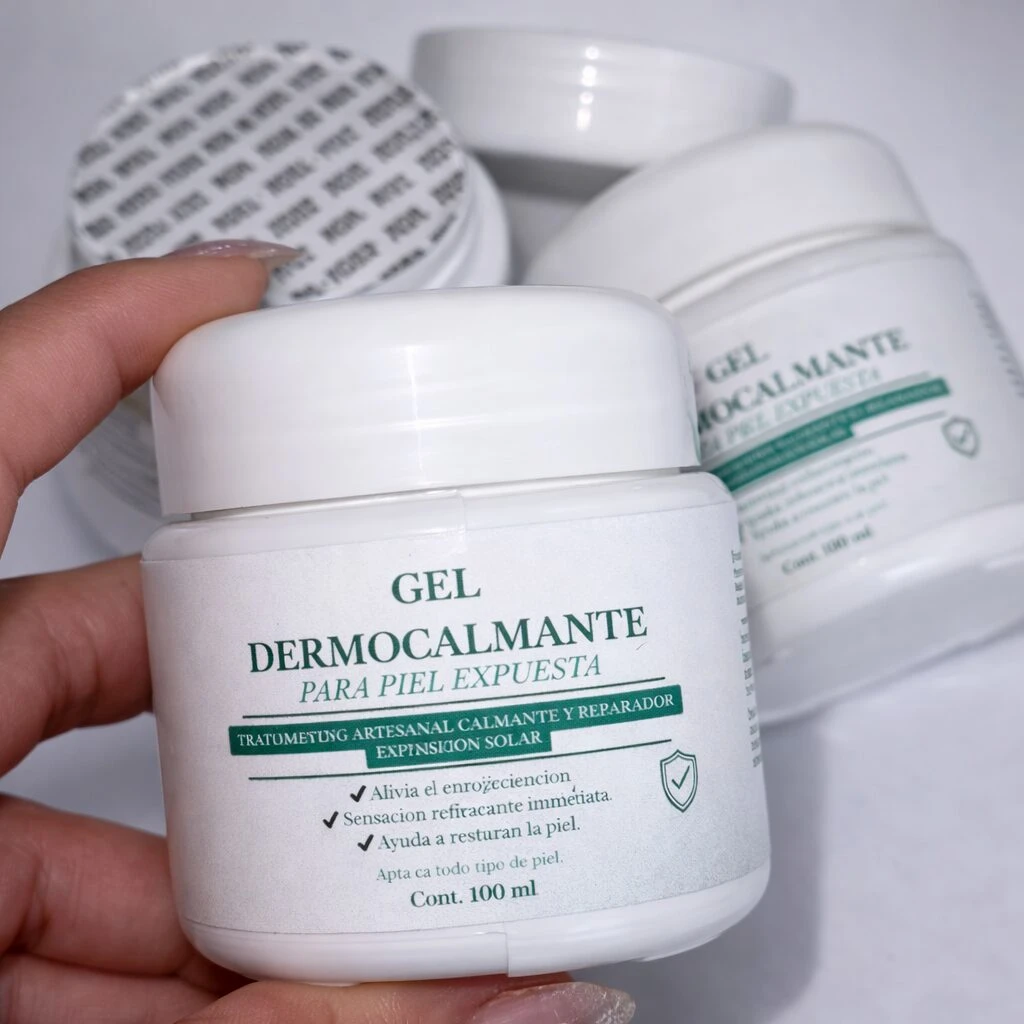 Gel Dermocalmante para piel expuesta, presentación de 100 ml en envase blanco con tapa rosca. Etiqueta verde y negra indica tratamiento artesanal calmante y reparador. Incluye dos unidades visibles en fondo, una parcialmente cubierta por la mano del usuario.