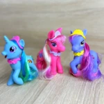 Tres figuras de pony de plástico con accesorios coloridos: uno azul con cuerno, otro rosa con peinado largo y el tercero morado con diadema amarilla, sobre superficie de madera clara.
