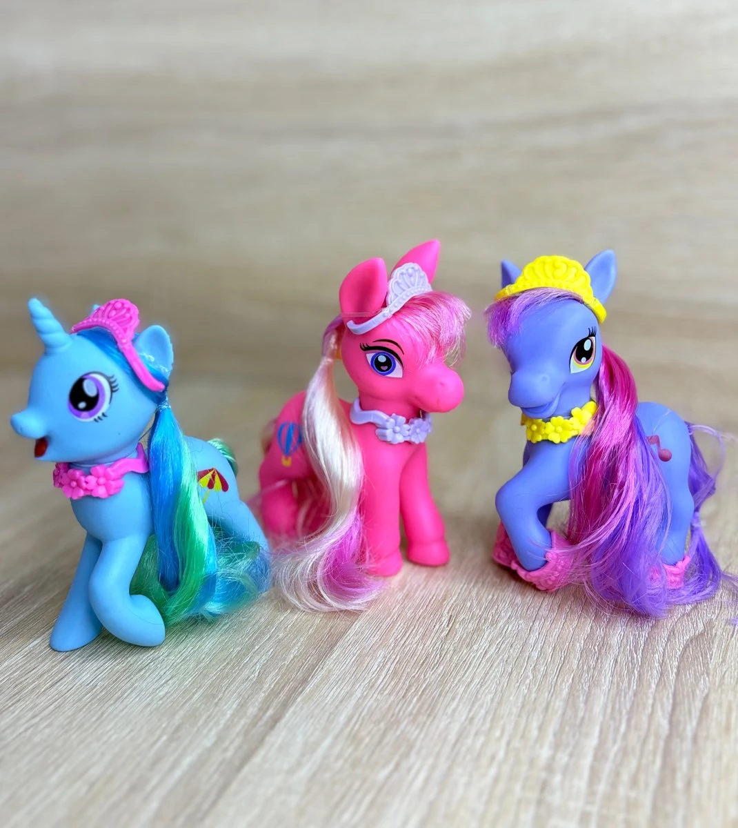 Tres figuras de pony de plástico con accesorios coloridos: uno azul con cuerno, otro rosa con peinado largo y el tercero morado con diadema amarilla, sobre superficie de madera clara.