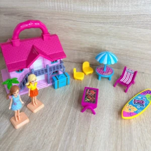 Casa de muñecas portátil rosa con accesorios de playa, incluyendo sombrilla, sillas, mesa, tabla de surf y dos figuras humanas sobre base.