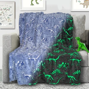 Manta de felpa con esqueletos de dinosaurios en azul y verde neón, colocada sobre sofá gris, ideal para niños.
