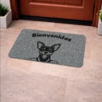 Tapete de entrada rectangular en color gris con textura granulada, diseño de perro chihuahua sonriente y texto "Bienvenidos" en negro, colocado frente a puerta de madera sobre piso de baldosas claras.