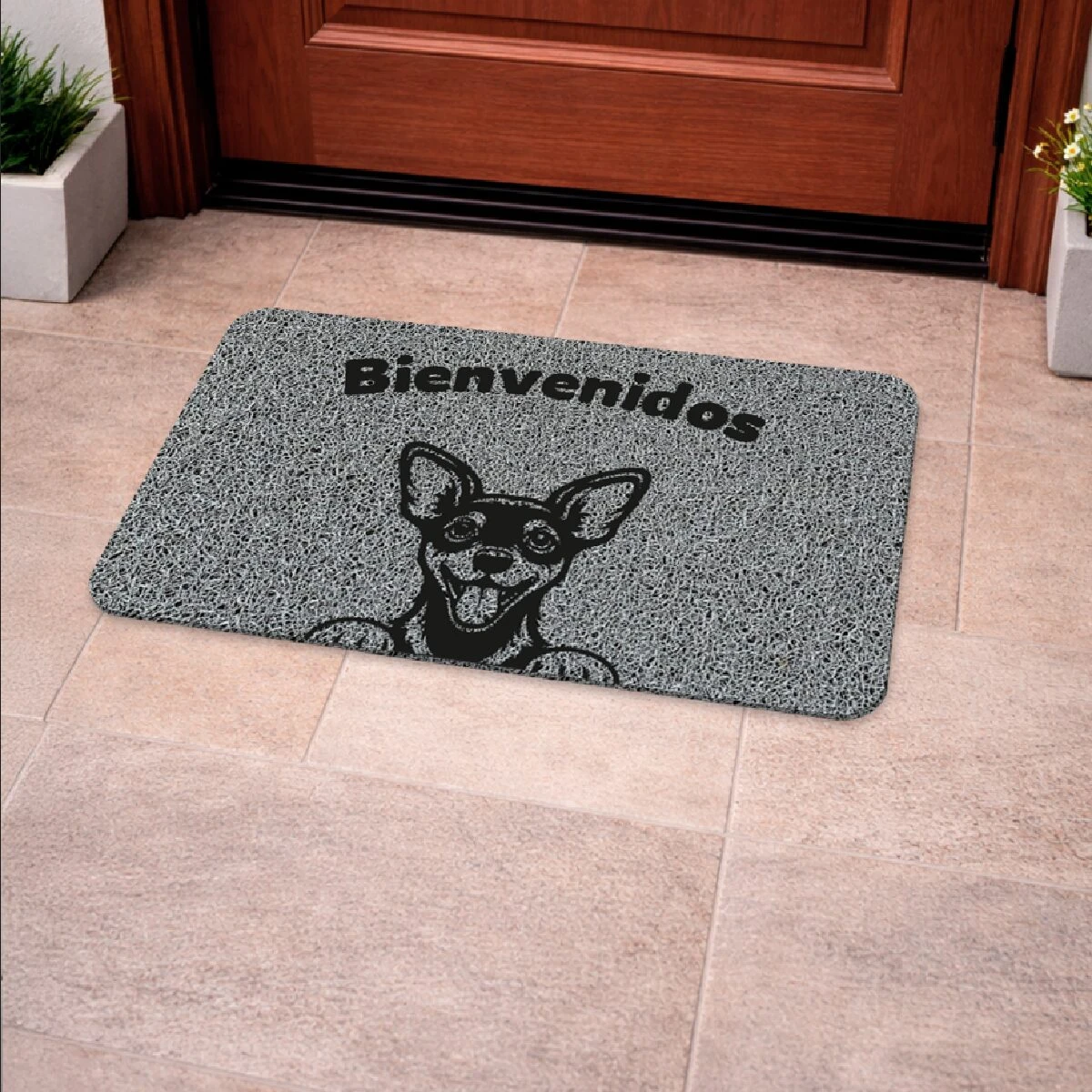 Tapete de entrada rectangular en color gris con textura granulada, diseño de perro chihuahua sonriente y texto "Bienvenidos" en negro, colocado frente a puerta de madera sobre piso de baldosas claras.