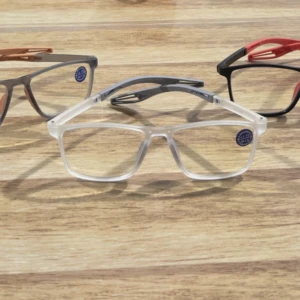 Tres pares de gafas de lectura TR90 con brazos flexibles, en colores gris, transparente y negro con detalles rojos, sobre superficie de madera.