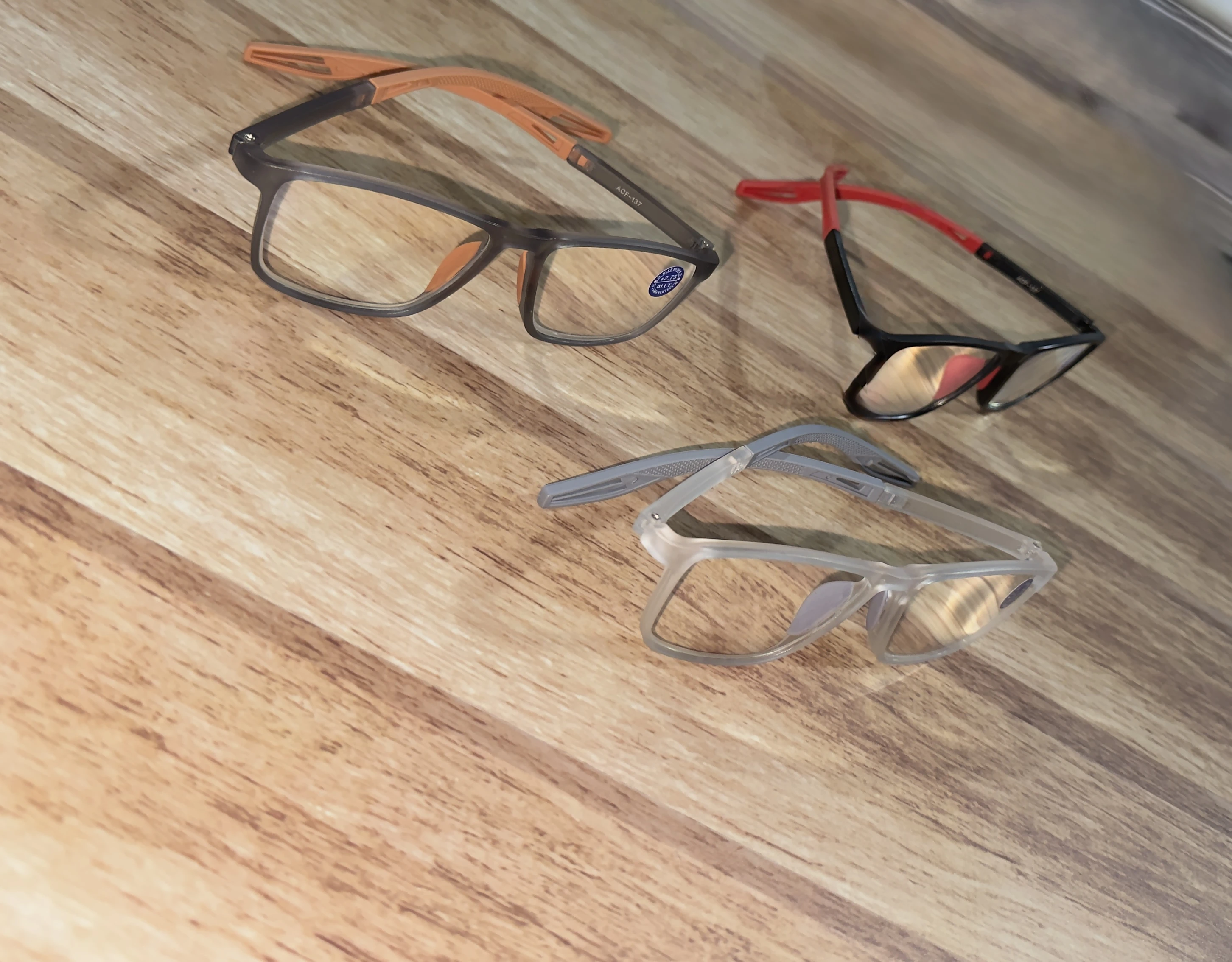 Tres pares de gafas de lectura con monturas en tonos gris, negro y transparente, sobre superficie de madera clara.