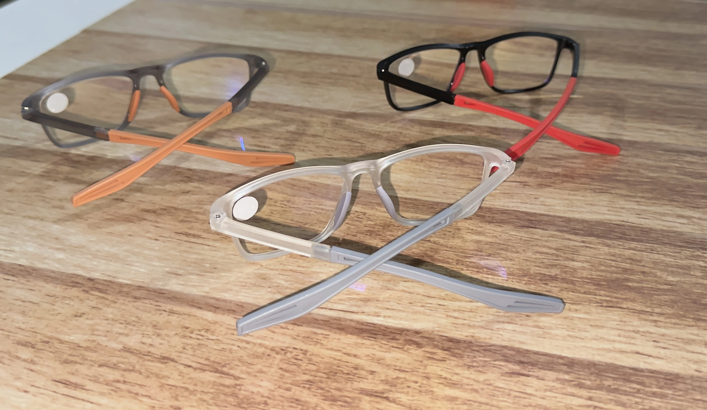 Tres pares de gafas de lectura con montura TR90 y brazos flexibles en colores gris, negro y transparente, dispuestas sobre superficie de madera.