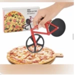 Cortador de pizza en forma de bicicleta roja con ruedas negras, sobre pizza de queso y jamón, con caja de presentación al fondo.