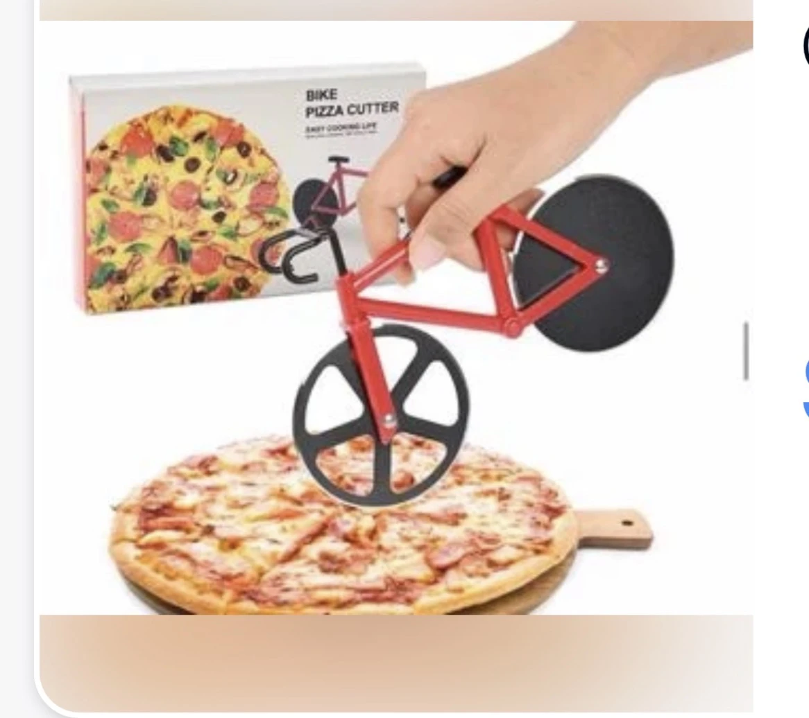 Cortador de pizza en forma de bicicleta roja con ruedas negras, sobre pizza de queso y jamón, con caja de presentación al fondo.