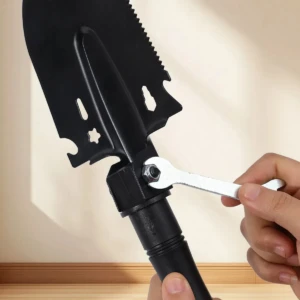 Pala plegable negra con sierra y brújula integrada, siendo ajustada con llave en mano, sobre fondo claro.