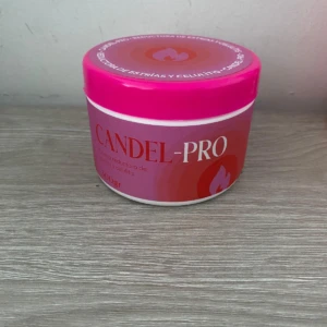 Contenedor redondo de crema Candel Pro en tono rosa con tapa brillante, sobre superficie de madera clara. Etiqueta frontal muestra nombre del producto y símbolo de llama.