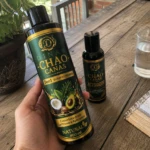 Botella de champú Chao Canas 5 en 1 con extracto de romero, color verde oscuro, etiqueta dorada y detalles de coco y aguacate, junto a un frasco más pequeño de activador, sobre mesa de madera con vaso de agua y planta al fondo.