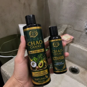 Champú Chao Canas 5 en 1 con extracto de romero, presentación de 400 ml, acompañado de su activador de 120 ml, ambos con diseño verde oscuro y detalles dorados, sostenidos en mano frente a un lavabo de concreto.