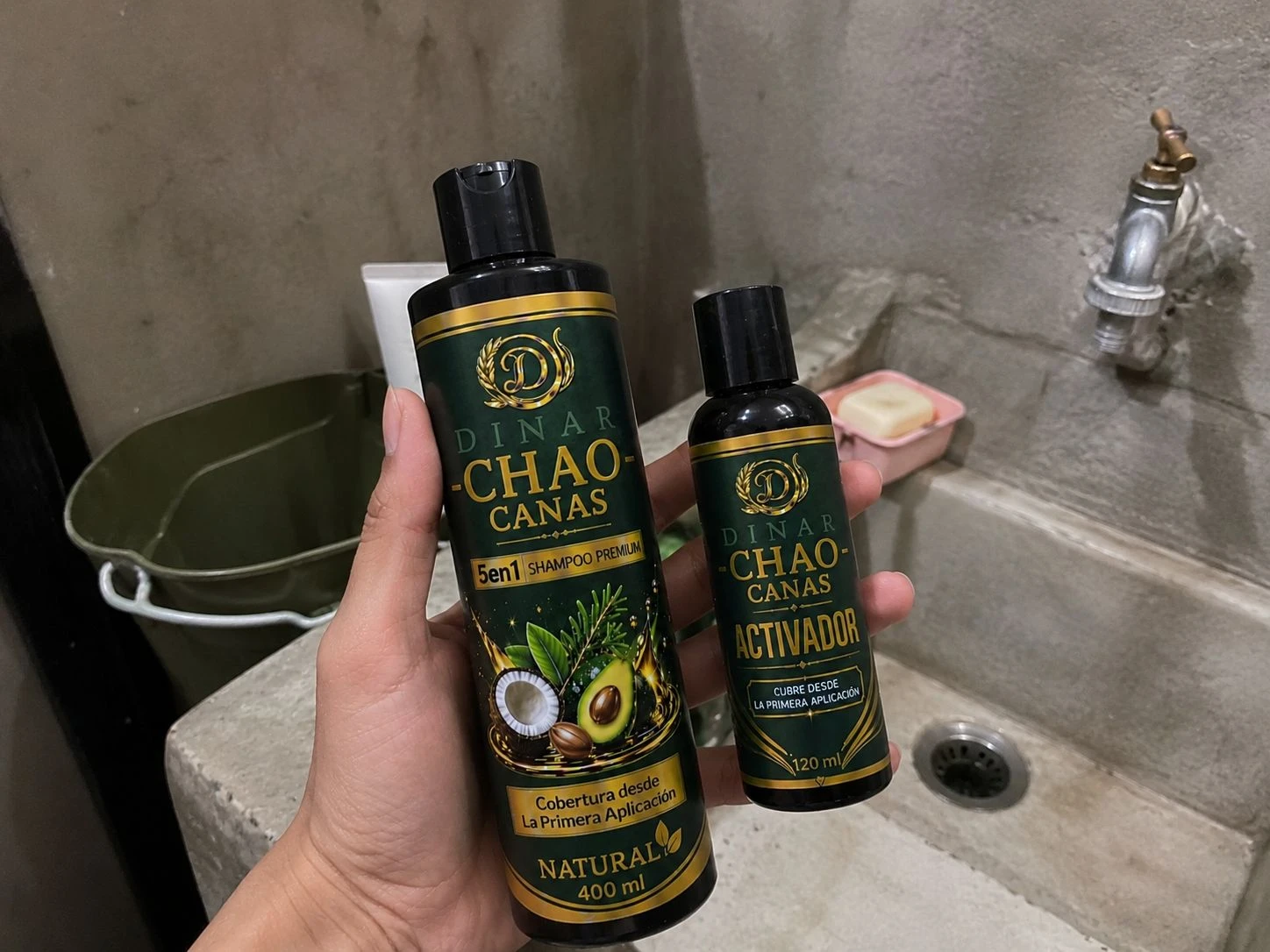 Champú Chao Canas 5 en 1 con extracto de romero, presentación de 400 ml, acompañado de su activador de 120 ml, ambos con diseño verde oscuro y detalles dorados, sostenidos en mano frente a un lavabo de concreto.