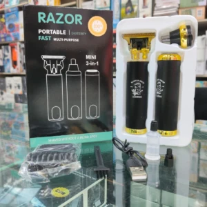 Pack de máquina eléctrica Razor híbrida negra con detalles dorados, incluye dos cabezales intercambiables, cargador USB y accesorios en caja de presentación sobre mostrador de vidrio.