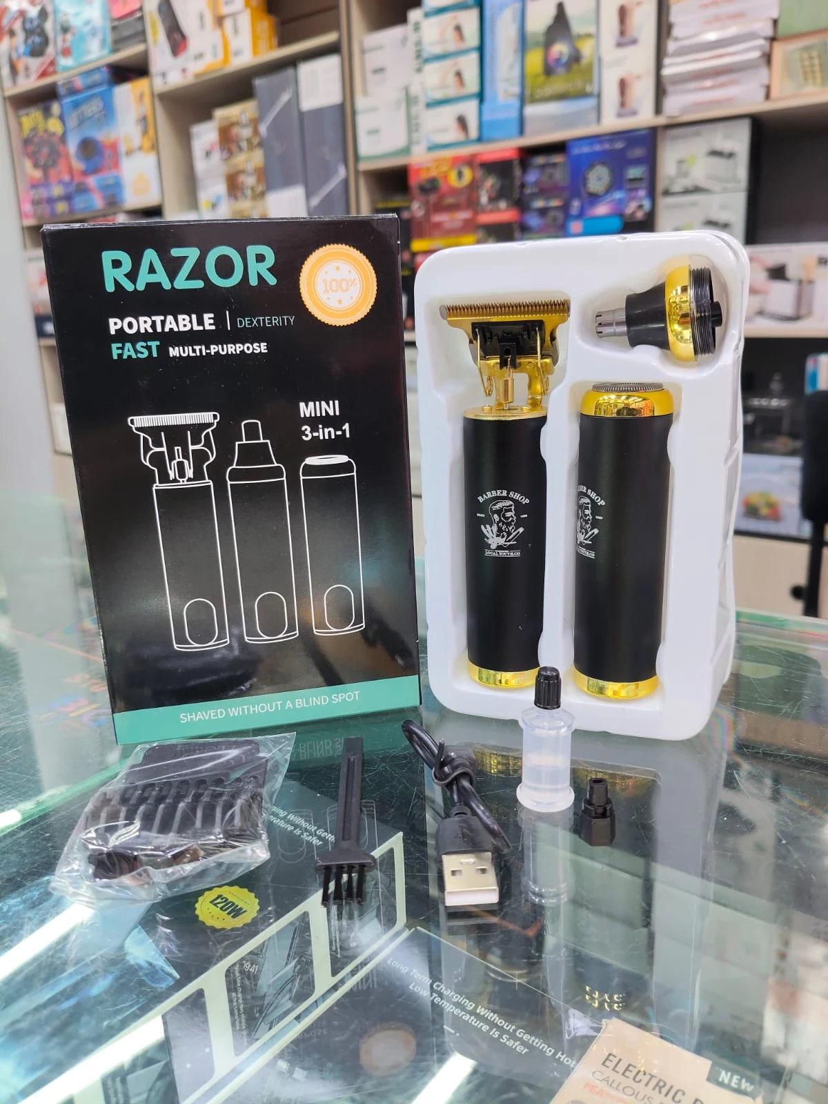Máquina eléctrica Razor híbrida negra y dorada con cabezales intercambiables, accesorios incluidos y caja de presentación sobre mostrador de vidrio.