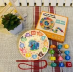 Reloj educativo de madera con bloques numerados y formas geométricas, caja naranja, cuerda roja y planta decorativa al fondo.