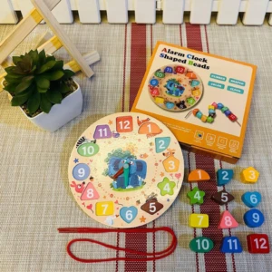 Reloj educativo de madera con bloques numerados y formas geométricas, caja naranja, cuerda roja y planta decorativa al fondo.