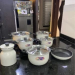 Bateria de cocina Alfusa White con jarra y sartén antiadherente, sobre encimera negra. Incluye ollas con tapas, sartén y jarra, todos en color blanco.
