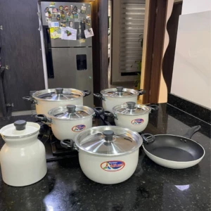 Bateria de cocina Alfusa White con jarra y sartén antiadherente, sobre encimera negra. Incluye ollas con tapas, sartén y jarra, todos en color blanco.