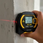 Cinta métrica láser negra y amarilla con pantalla LCD mostrando medidas en metros, emitiendo rayo láser rojo sobre superficie de concreto, usada para medición precisa en construcción.