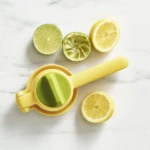 Exprimidor manual de cítricos en color amarillo, con mango ergonómico y base circular verde. A su alrededor, rodajas de limón y lima, una de ellas ya exprimida, sobre superficie blanca con vetas grises.
