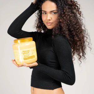 Mujer con rizos oscuros sosteniendo un frasco amarillo de tratamiento capilar Honey Plus, con miel y proteínas, sobre fondo gris neutro.