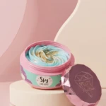Jarrín cosmético abierto con crema azul y dorada, tapa rosa brillante al lado, sobre pedestal blanco en fondo rosa.
