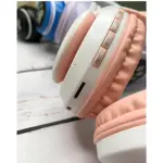 Auriculares diadema en rosa pastel y blanco, mostrando controles laterales con botones táctiles y puerto USB-C, sobre superficie blanca con otros accesorios al fondo.