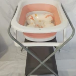 Soporte metálico plegable para bañera de bebé, color blanco con base rosa, incluye tina con accesorios de baño visibles.