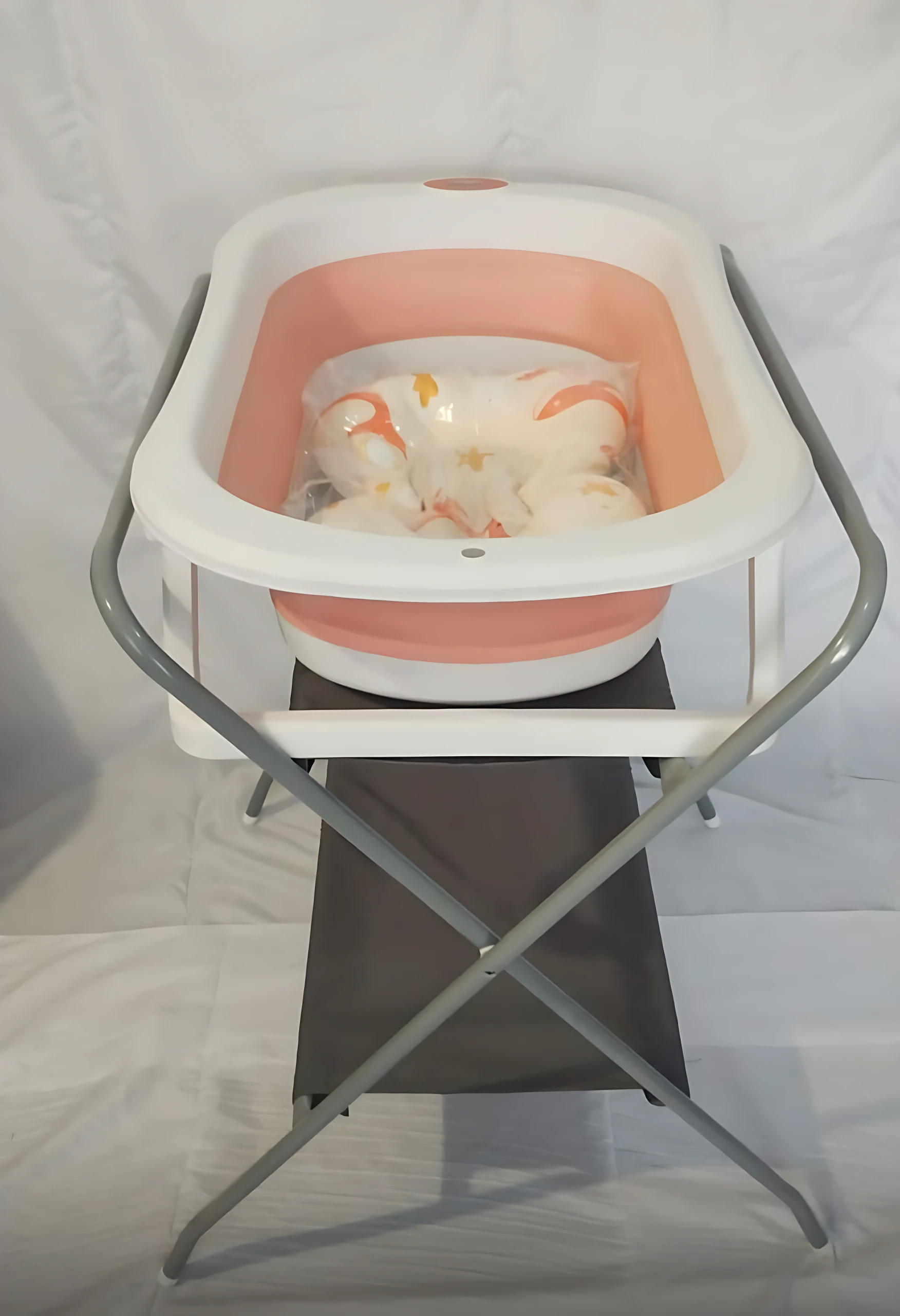 Soporte metálico plegable para bañera de bebé, color blanco con base rosa, incluye tina con accesorios de baño visibles.