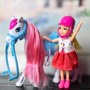 Barbie niña con casco rosa montando caballo de peluche blanco y crin rosa, sobre superficie oscura.