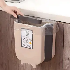 Basurera portátil beige con tapa negra, montada en puerta de cocina mediante gancho superior; incluye bolsa plástica transparente y etiqueta informativa frontal.