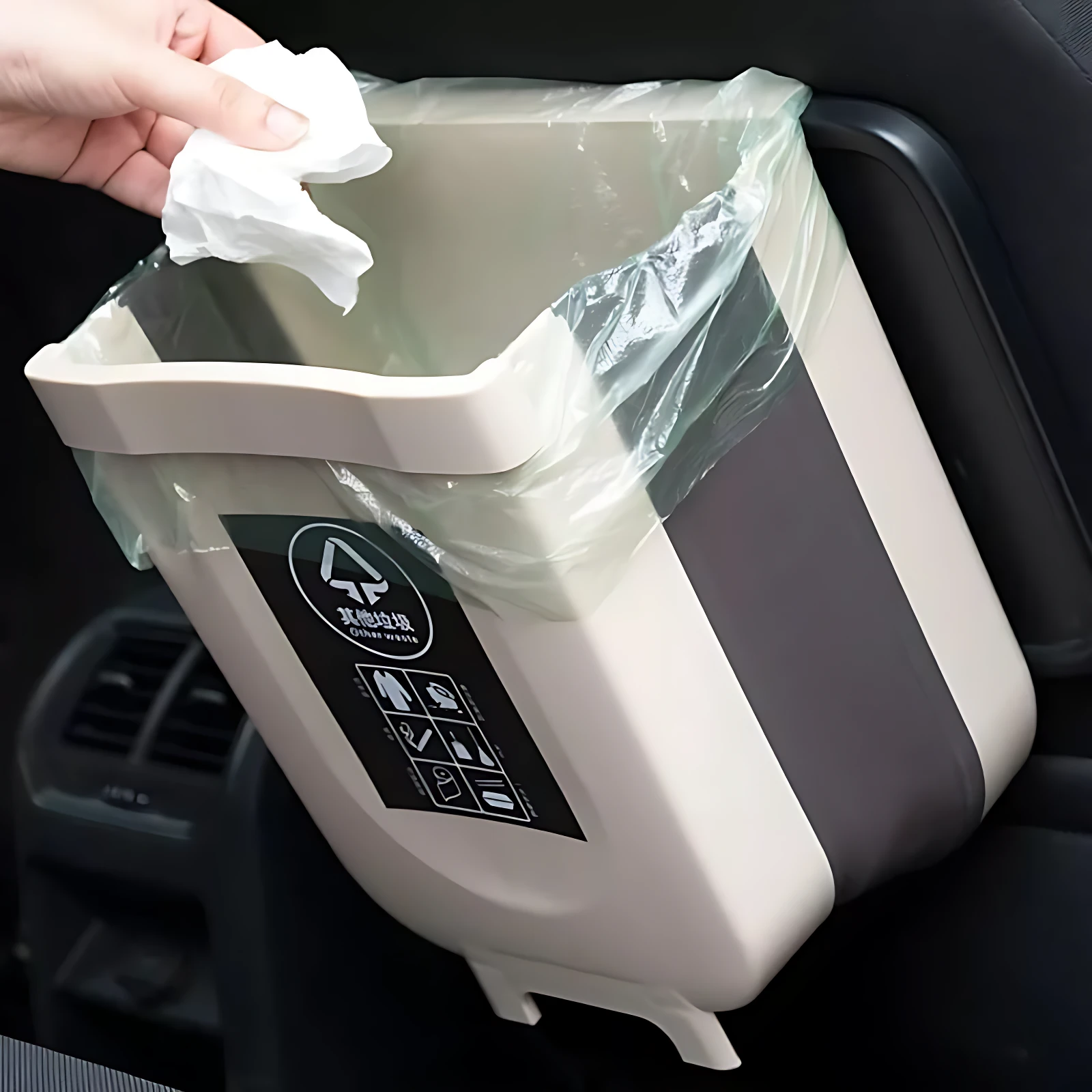 Basurera portátil plegable beige con bolsa plástica transparente, siendo usada en interior de vehículo, con etiqueta negra frontal y gancho lateral visible.