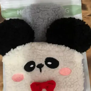 Bolsa de felpa suave con diseño de panda, orejas negras, mejillas rosadas y lazo rojo, presentada en su empaque original.