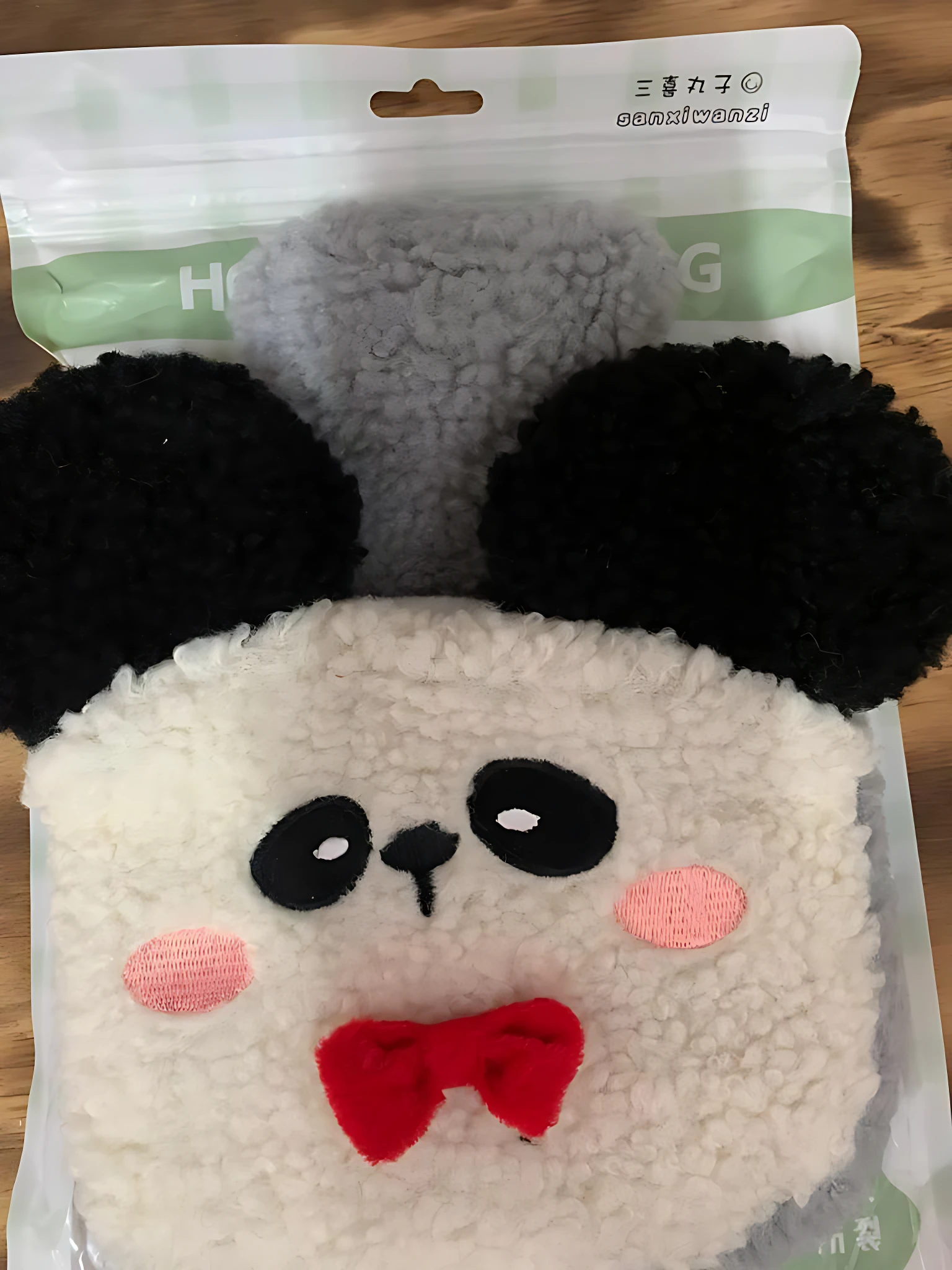 Bolsa de felpa suave con diseño de panda, orejas negras, mejillas rosadas y lazo rojo, presentada en su empaque original.